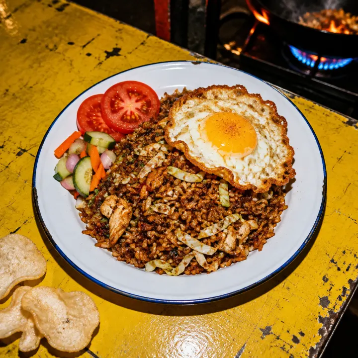 Nasi goreng kecap manis gelap dengan kol, potongan ayam, telur ceplok, irisan tomat, dan acar di piring enamel.